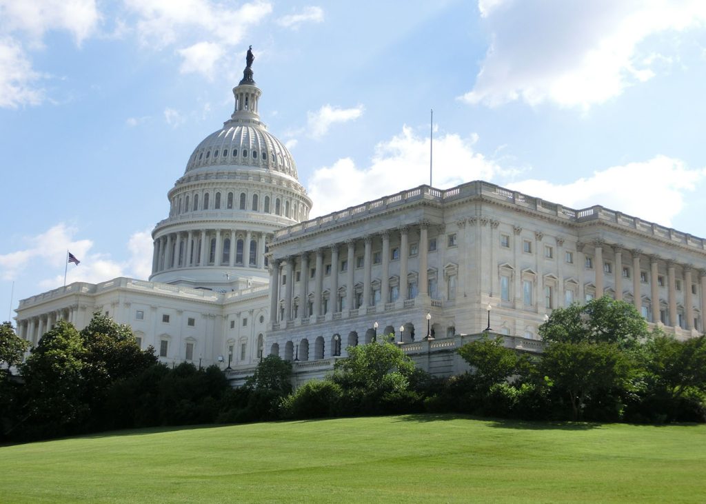 The United States Capitol Building on a sunny day, with a clear blue sky and green lawns in the foreground.