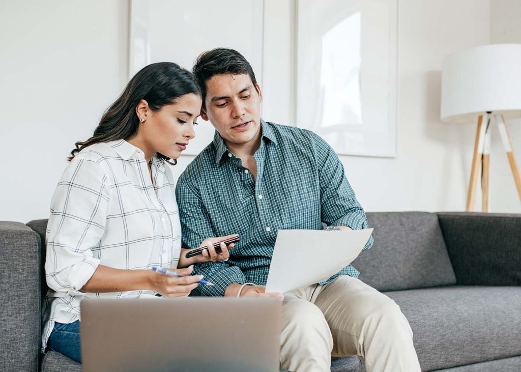A couple is sitting on a sofa, engaged in reviewing documents and a laptop, with a smartphone in hand, suggesting a collaborative financial planning or discussion at home.