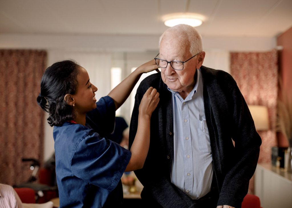 A caregiver in blue scrubs kindly helps an elderly man with glasses put on his jacket, wearing a blue shirt and a black cardigan.