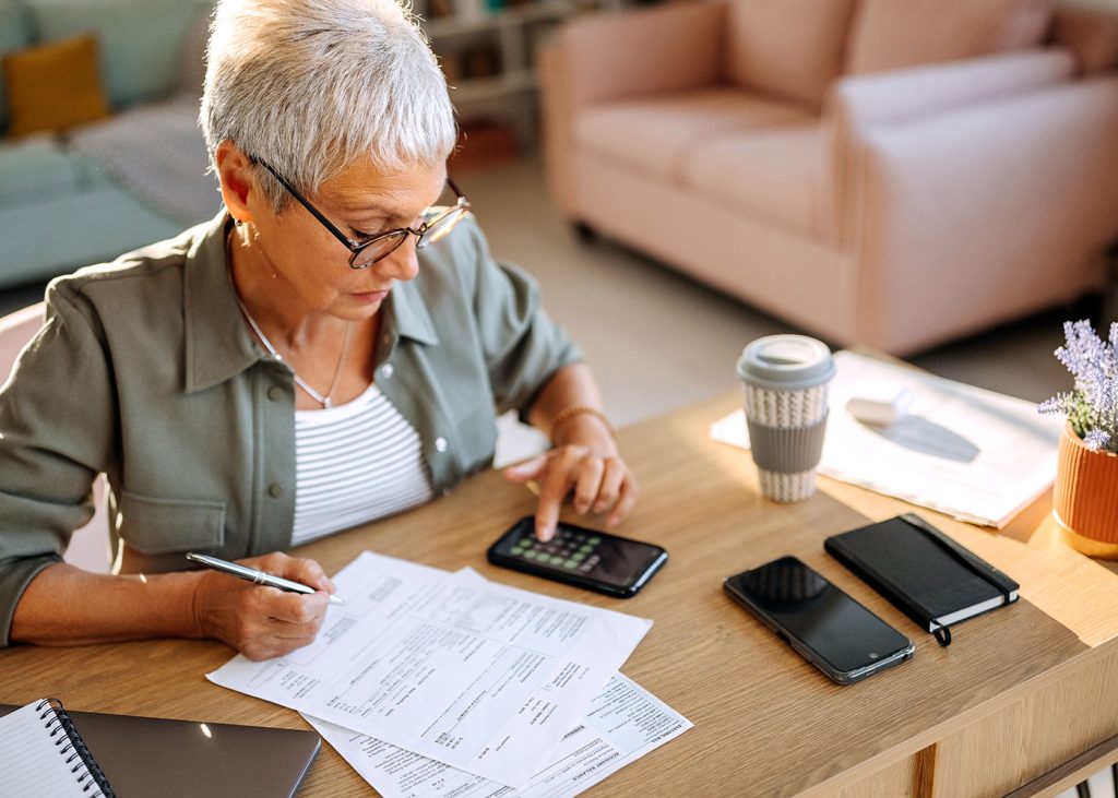 A senior woman focused on doing taxes at home, surrounded by paperwork and using a calculator.