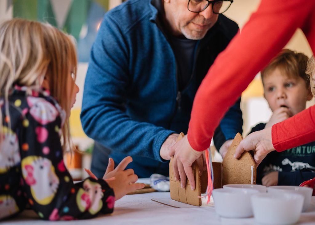 A man and woman assisting children in constructing gingerbread houses during a holiday event.