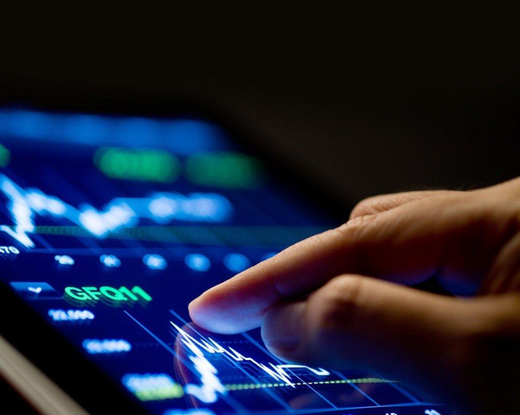 A hand with fingertips on a tablet screen displaying a stock market graph.